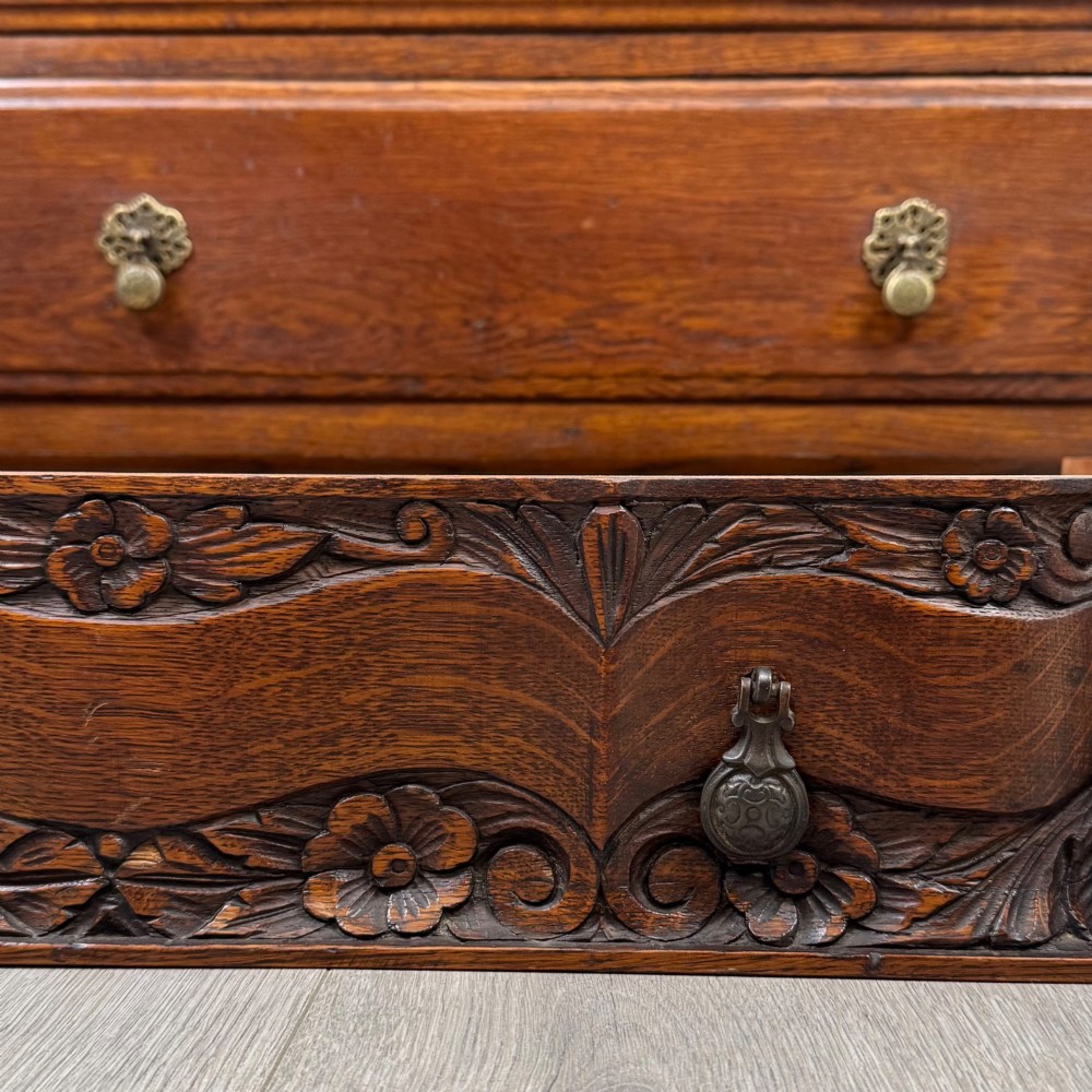A dark carved oak chest of drawers featuring three drawers, floral relief carvings, and brass pendant handles set on carved bracket feet. View XI
