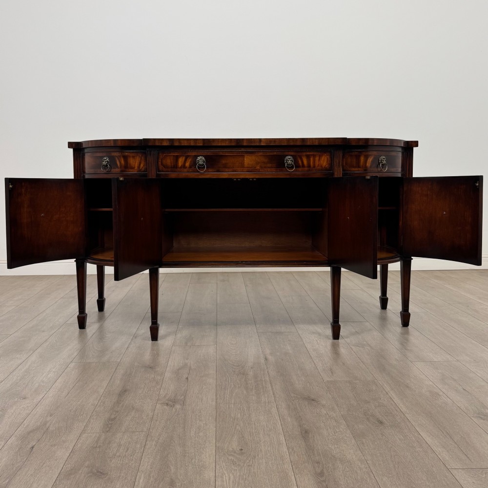 A 20th-century flame mahogany sideboard featuring a bow front design, drawers, cupboard doors, brass lion mask ring handles, and square tapered legs. View II