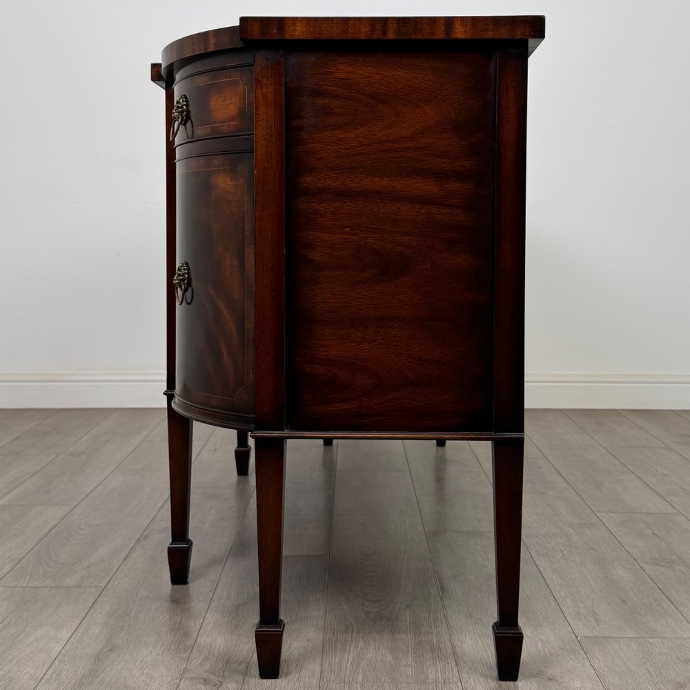 A 20th-century flame mahogany sideboard featuring a bow front design, drawers, cupboard doors, brass lion mask ring handles, and square tapered legs. View IV