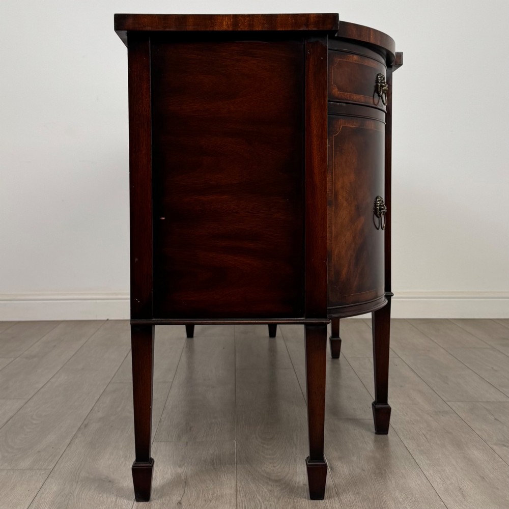 A 20th-century flame mahogany sideboard featuring a bow front design, drawers, cupboard doors, brass lion mask ring handles, and square tapered legs. View VIII