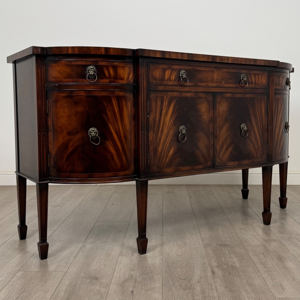 A 20th-century flame mahogany sideboard featuring a bow front design, drawers, cupboard doors, brass lion mask ring handles, and square tapered legs. View IX