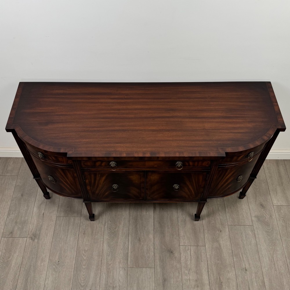 A 20th-century flame mahogany sideboard featuring a bow front design, drawers, cupboard doors, brass lion mask ring handles, and square tapered legs. View X