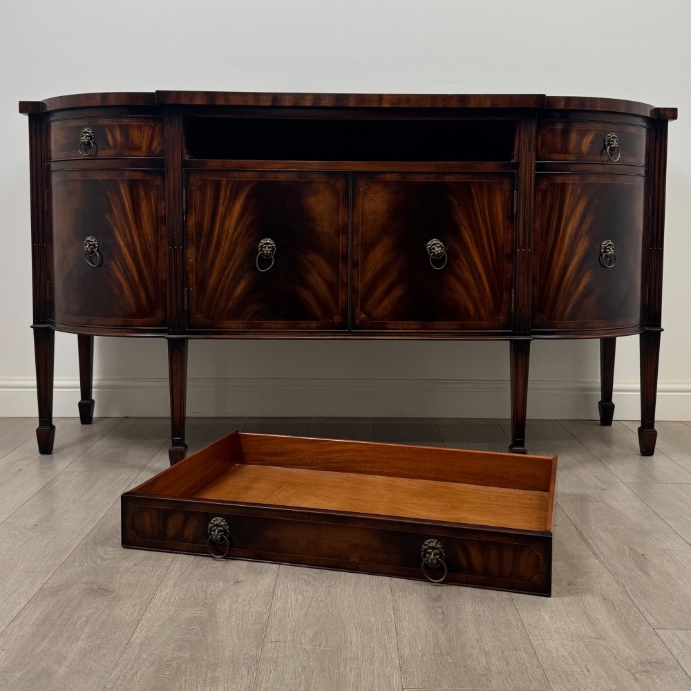 A 20th-century flame mahogany sideboard featuring a bow front design, drawers, cupboard doors, brass lion mask ring handles, and square tapered legs. View XI