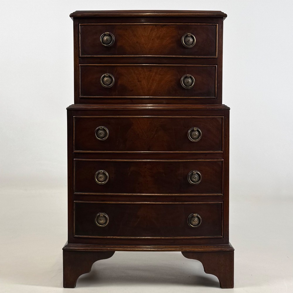 A small dark mahogany chest of drawers with five stacked drawers, brass ring handles, and bracket feet.