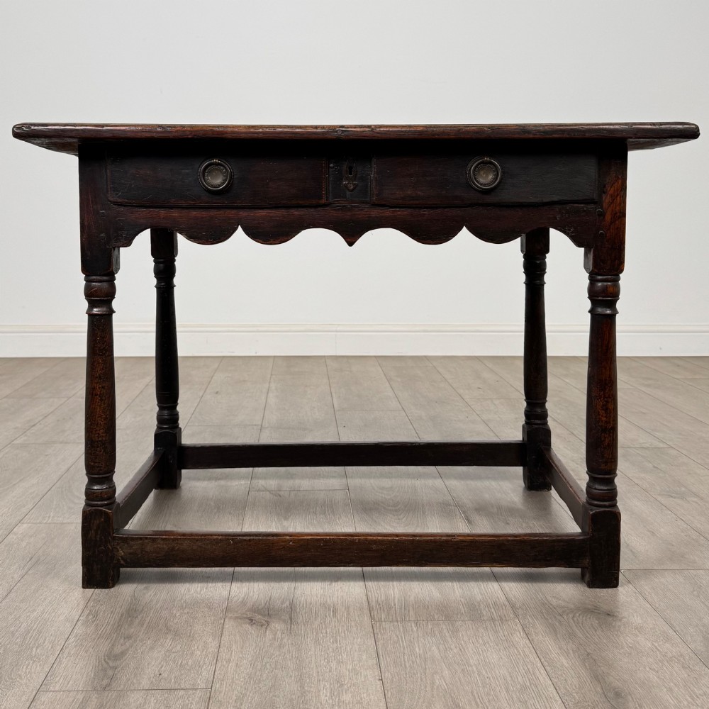 An 18th-century dark oak side table featuring turned legs, a scalloped apron frieze, a single drawer with brass hardware, and perimeter stretchers.