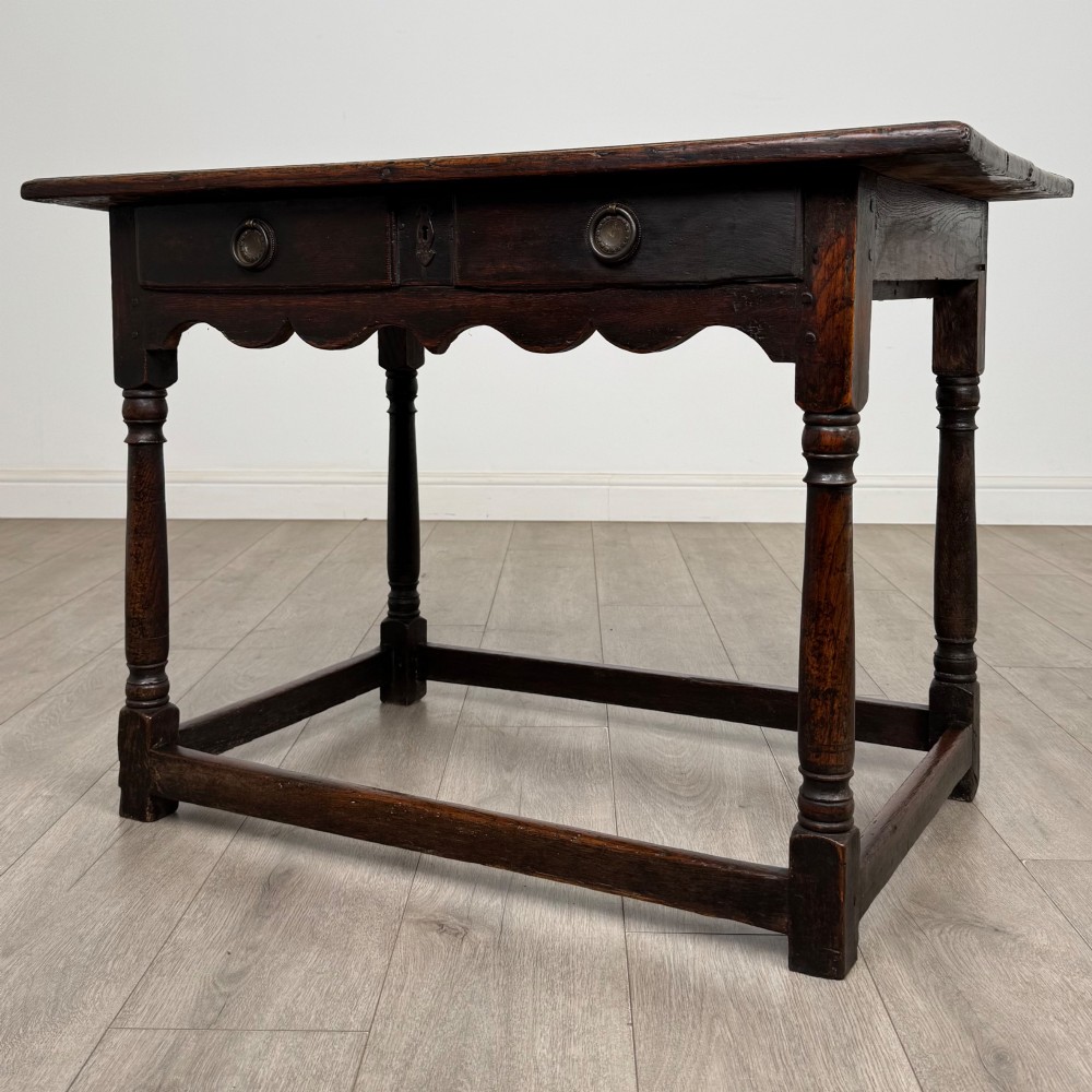 An 18th-century dark oak side table featuring turned legs, a scalloped apron frieze, a single drawer with brass hardware, and perimeter stretchers. View II