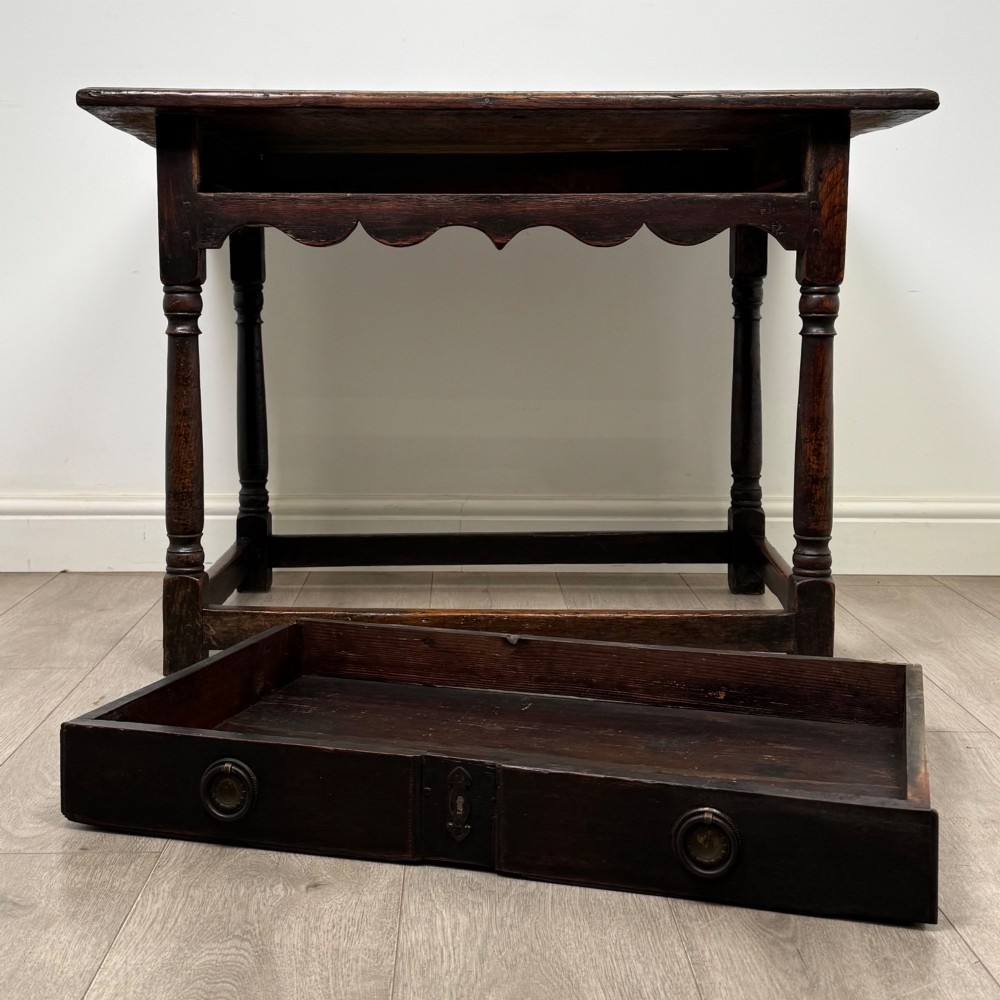 An 18th-century dark oak side table featuring turned legs, a scalloped apron frieze, a single drawer with brass hardware, and perimeter stretchers. View X