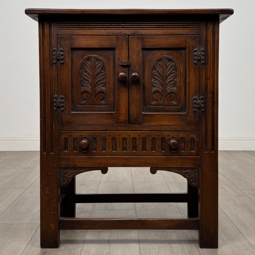 A dark brown carved oak cupboard featuring two arched leaf panel doors, a fluted frieze drawer, and wooden knobs set on squared legs.