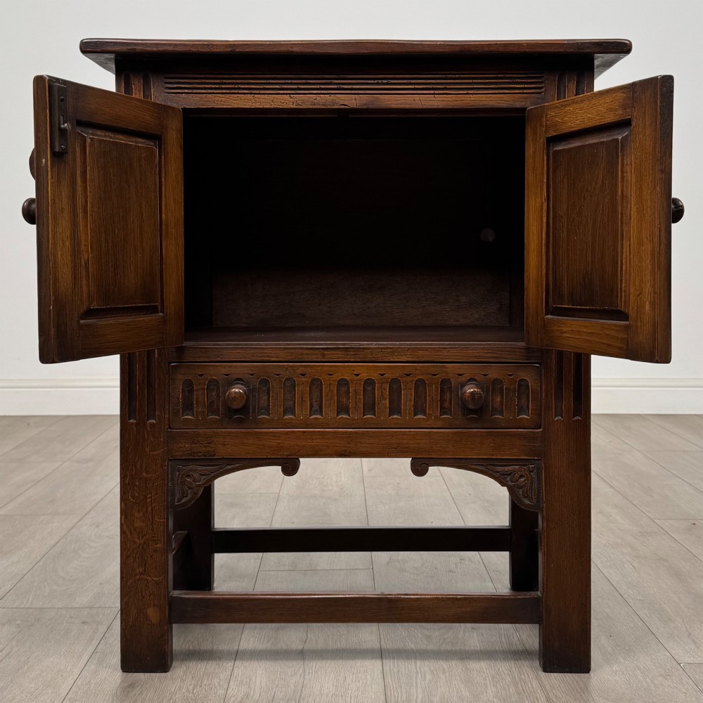 A dark brown carved oak cupboard featuring two arched leaf panel doors, a fluted frieze drawer, and wooden knobs set on squared legs. View II