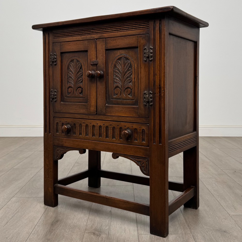 A dark brown carved oak cupboard featuring two arched leaf panel doors, a fluted frieze drawer, and wooden knobs set on squared legs. View III