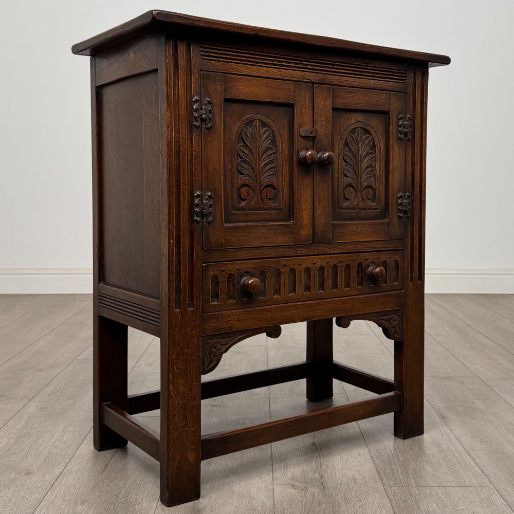 A dark brown carved oak cupboard featuring two arched leaf panel doors, a fluted frieze drawer, and wooden knobs set on squared legs. View IX