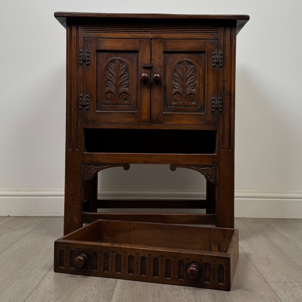 A dark brown carved oak cupboard featuring two arched leaf panel doors, a fluted frieze drawer, and wooden knobs set on squared legs. View XI