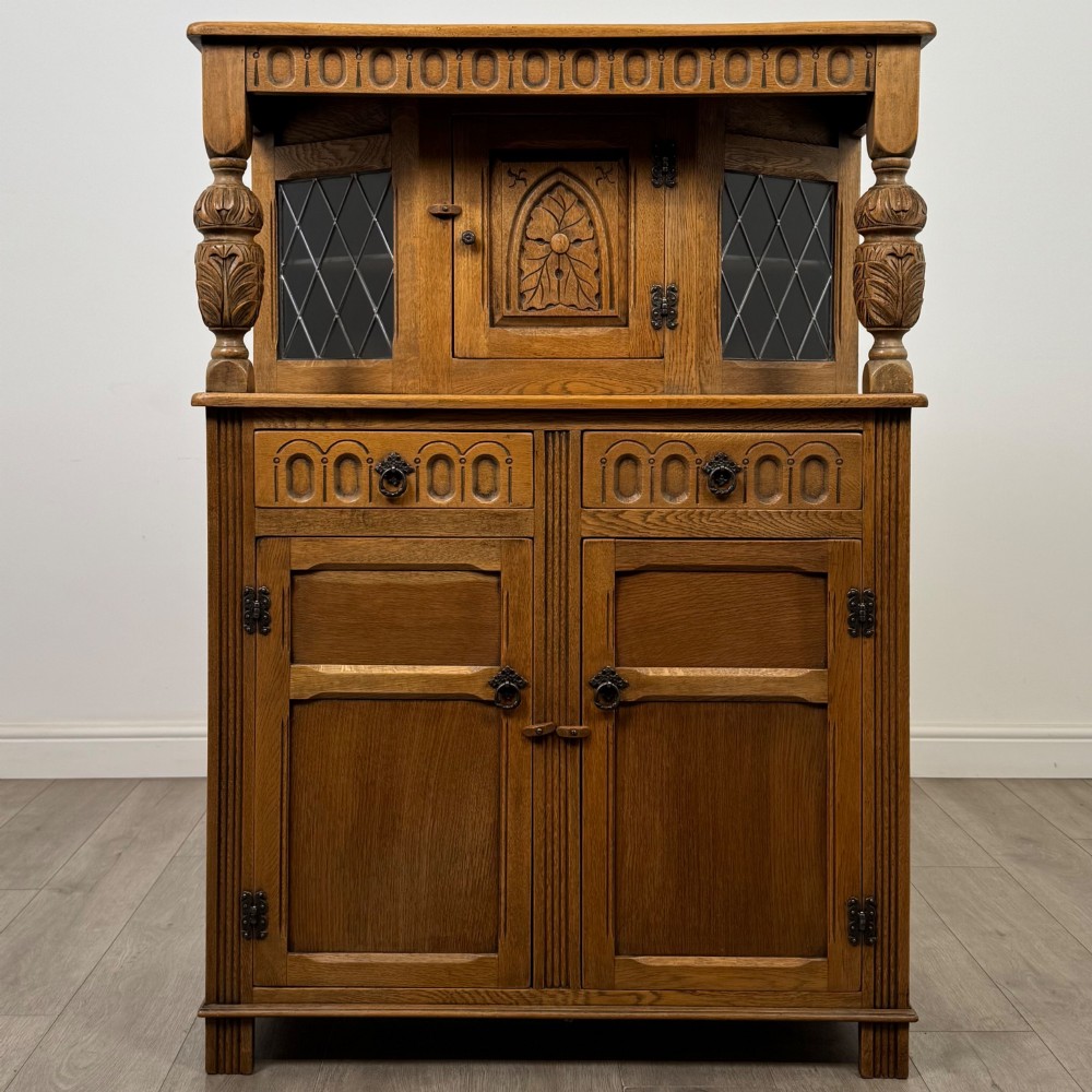 A twentieth-century golden oak court cupboard featuring carved panels, leaded glass side doors, and turned bulbous supports.