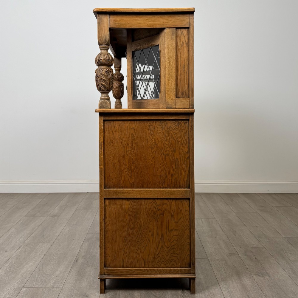 A twentieth-century golden oak court cupboard featuring carved panels, leaded glass side doors, and turned bulbous supports. View IV