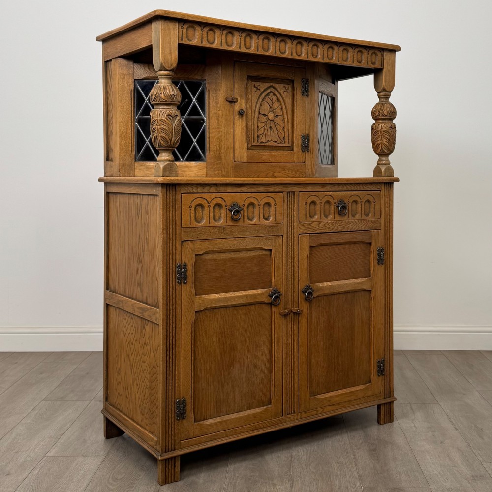 A twentieth-century golden oak court cupboard featuring carved panels, leaded glass side doors, and turned bulbous supports. View IX