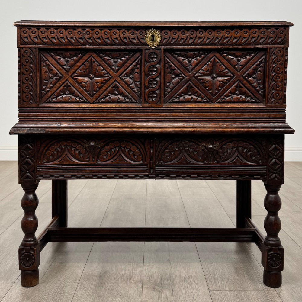 A 19th-century carved dark oak chest on stand featuring geometric relief panels, brass keyhole escutcheon, lower drawers, and turned legs.
