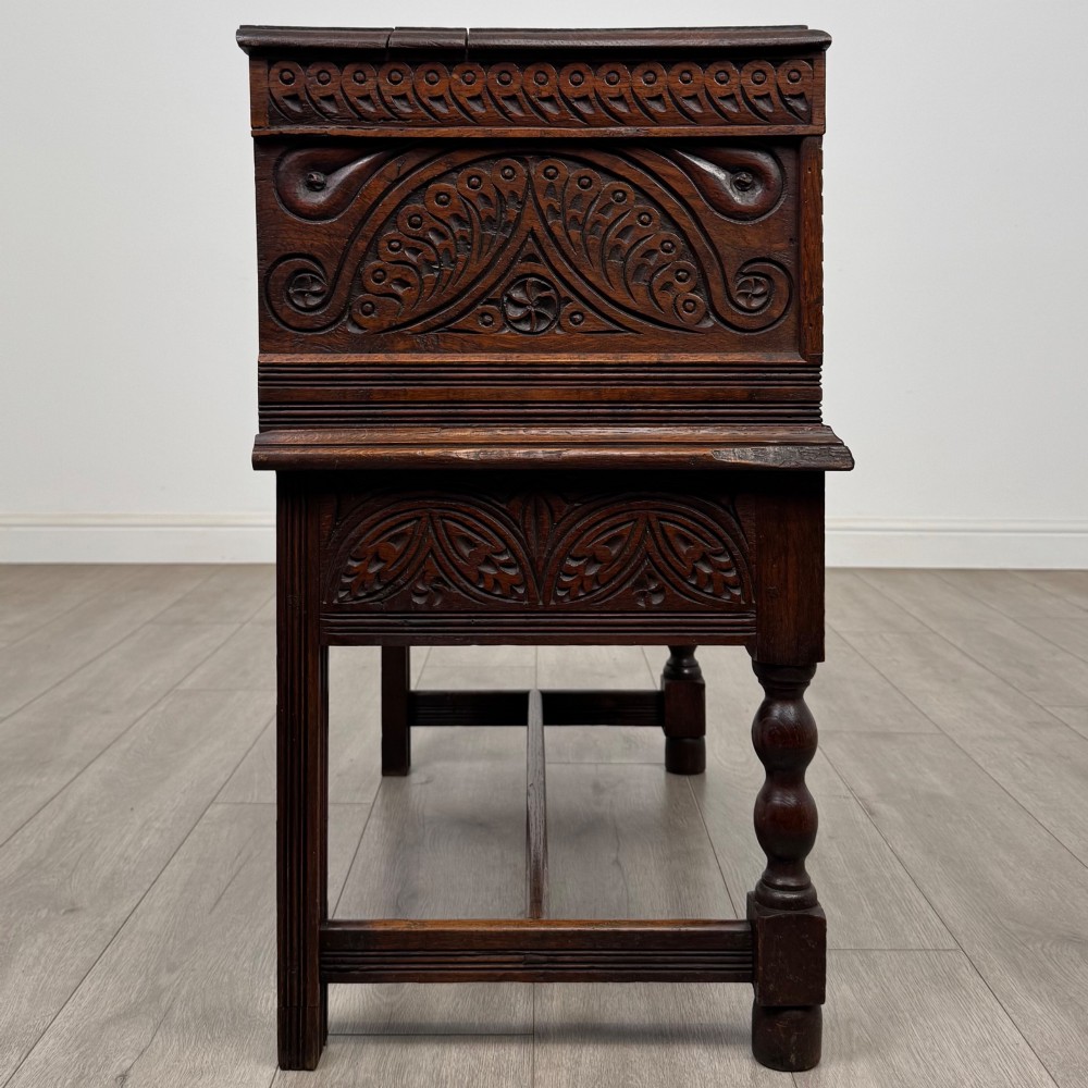A 19th-century carved dark oak chest on stand featuring geometric relief panels, brass keyhole escutcheon, lower drawers, and turned legs. View VII