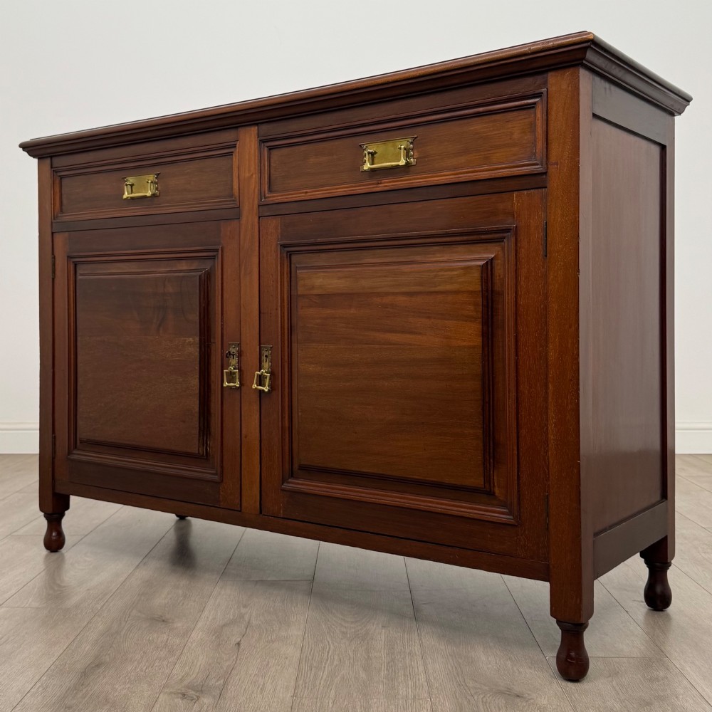 An antique Edwardian mahogany sideboard featuring two top drawers with brass drop handles above two field-panelled cupboard doors, resting on short turned feet. View III