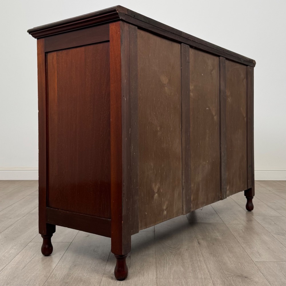 An antique Edwardian mahogany sideboard featuring two top drawers with brass drop handles above two field-panelled cupboard doors, resting on short turned feet. View V