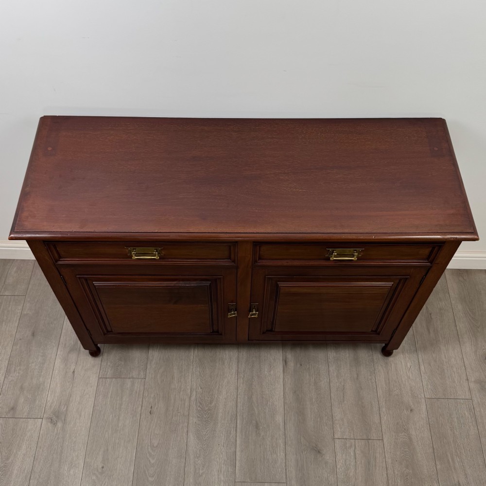 An antique Edwardian mahogany sideboard featuring two top drawers with brass drop handles above two field-panelled cupboard doors, resting on short turned feet. View X