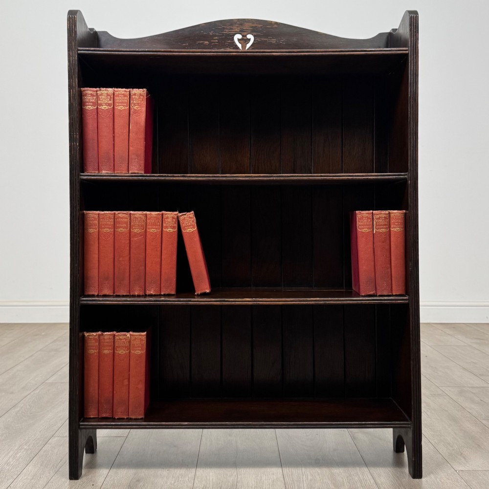 A dark brown oak open bookcase featuring three tiers, vertical plank backboard, and a shaped top rail with a central cut-out motif.