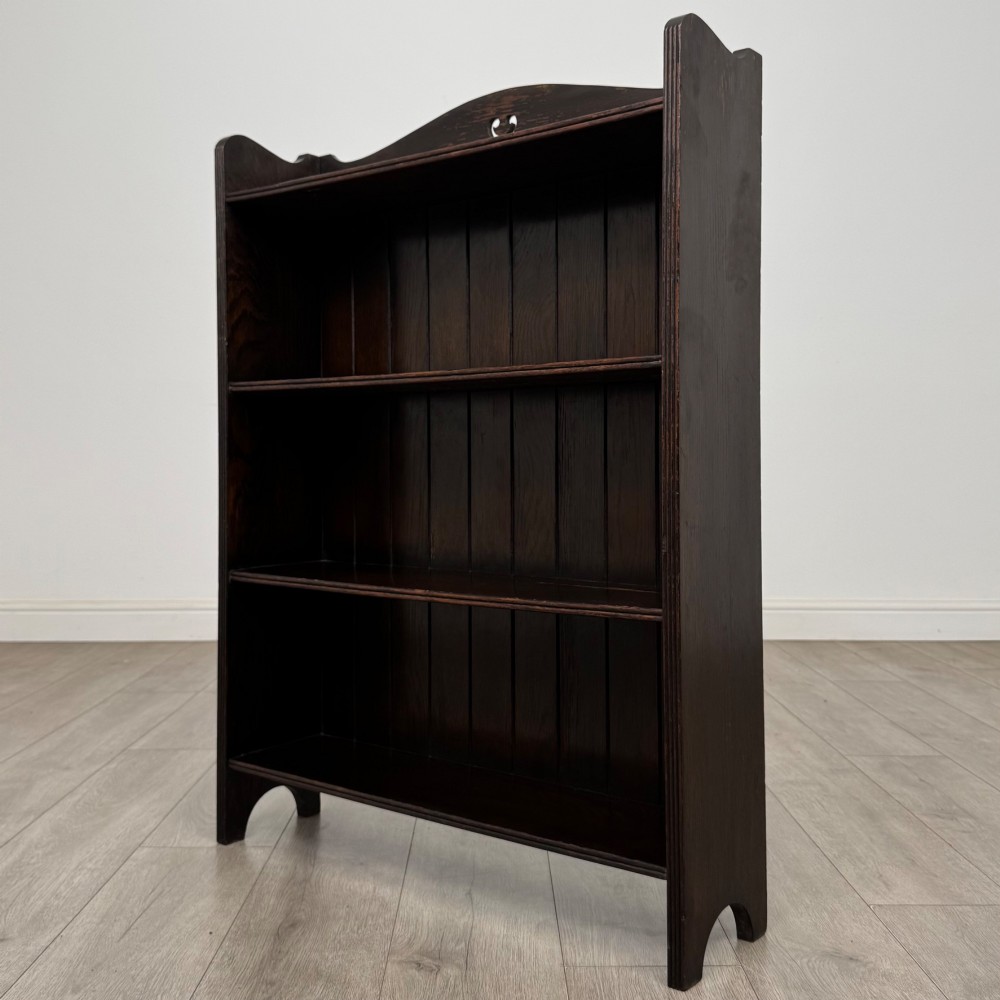 A dark brown oak open bookcase featuring three tiers, vertical plank backboard, and a shaped top rail with a central cut-out motif. View III