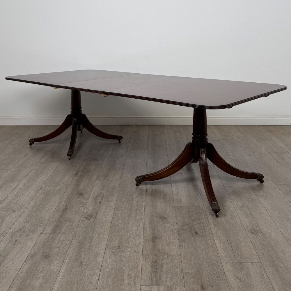 A 20th-century mahogany twin pedestal dining table featuring an extension leaf, reeded edges, and splayed quadruped feet with brass castors.