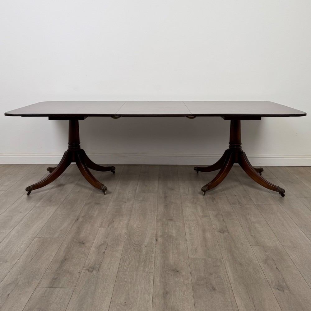A 20th-century mahogany twin pedestal dining table featuring an extension leaf, reeded edges, and splayed quadruped feet with brass castors. View II
