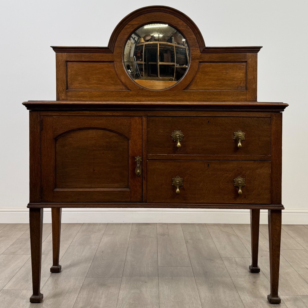 An Edwardian oak sideboard featuring a raised gallery back with a central circular mirror, a left cupboard door, two right drawers, and brass drop handles.