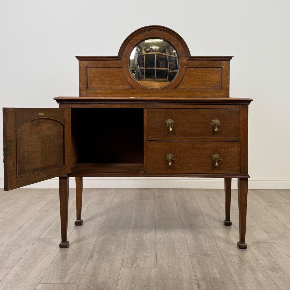 An Edwardian oak sideboard featuring a raised gallery back with a central circular mirror, a left cupboard door, two right drawers, and brass drop handles. View II