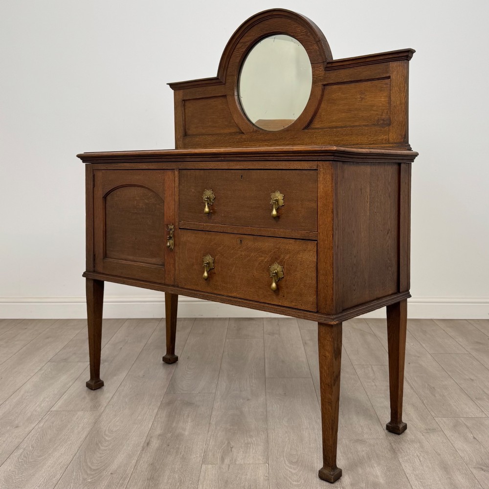An Edwardian oak sideboard featuring a raised gallery back with a central circular mirror, a left cupboard door, two right drawers, and brass drop handles. View III