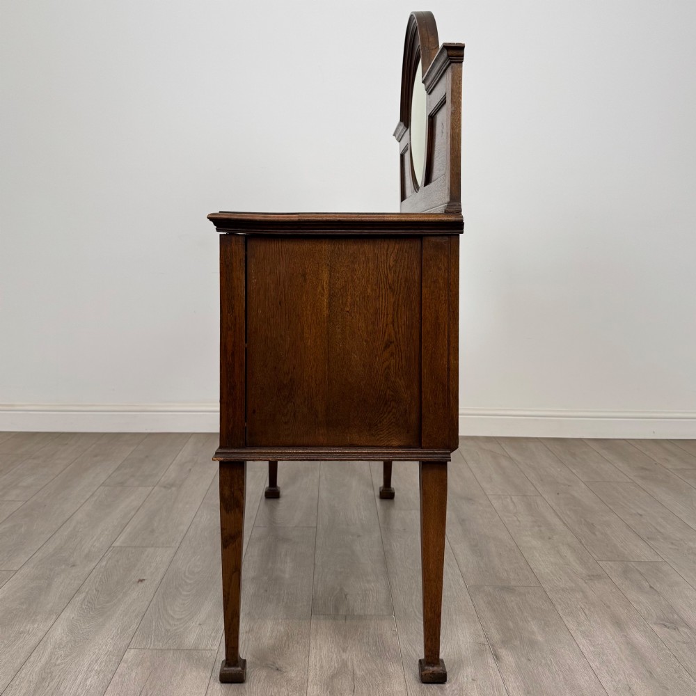 An Edwardian oak sideboard featuring a raised gallery back with a central circular mirror, a left cupboard door, two right drawers, and brass drop handles. View IV