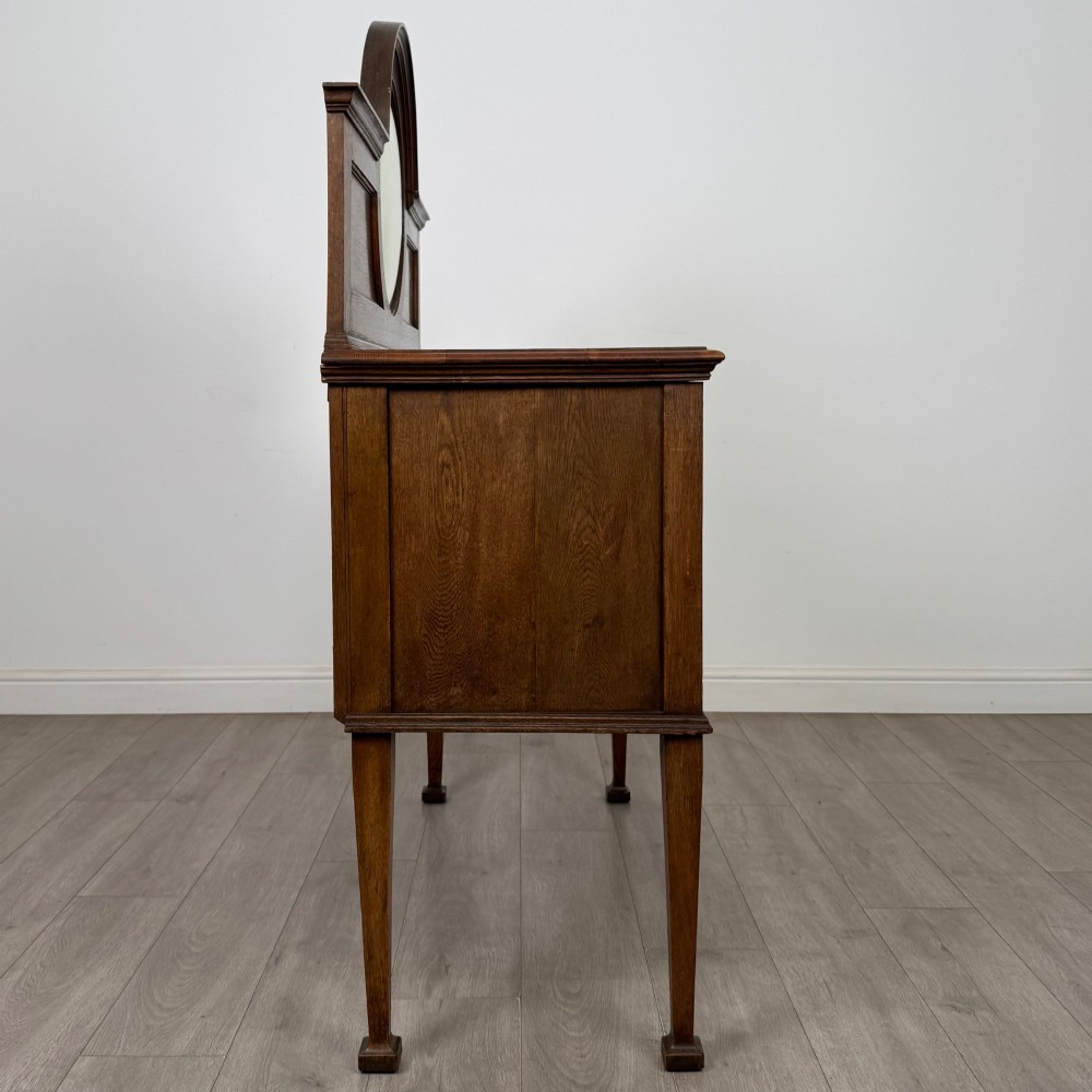 An Edwardian oak sideboard featuring a raised gallery back with a central circular mirror, a left cupboard door, two right drawers, and brass drop handles. View VIII