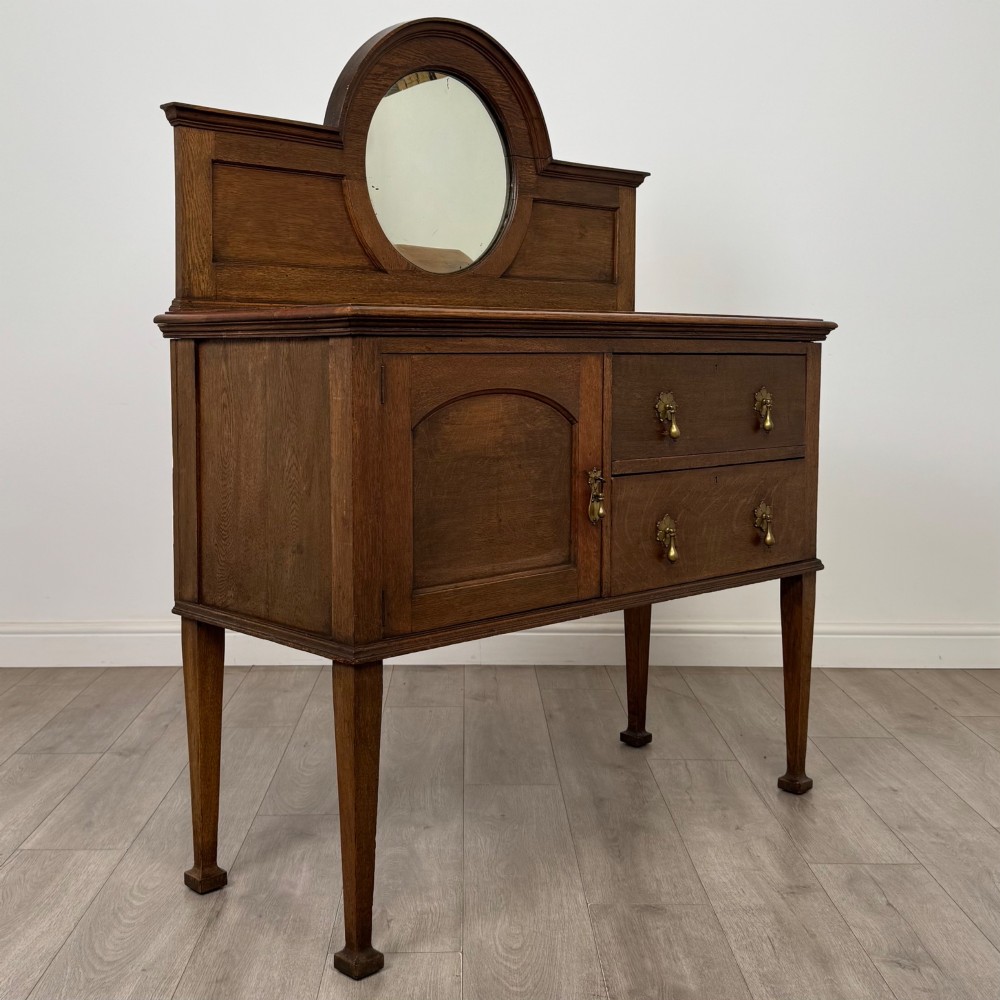 An Edwardian oak sideboard featuring a raised gallery back with a central circular mirror, a left cupboard door, two right drawers, and brass drop handles. View IX