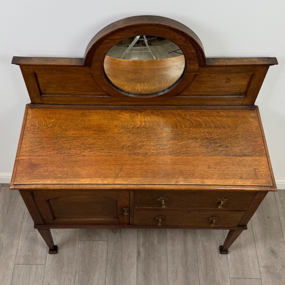 An Edwardian oak sideboard featuring a raised gallery back with a central circular mirror, a left cupboard door, two right drawers, and brass drop handles. View X