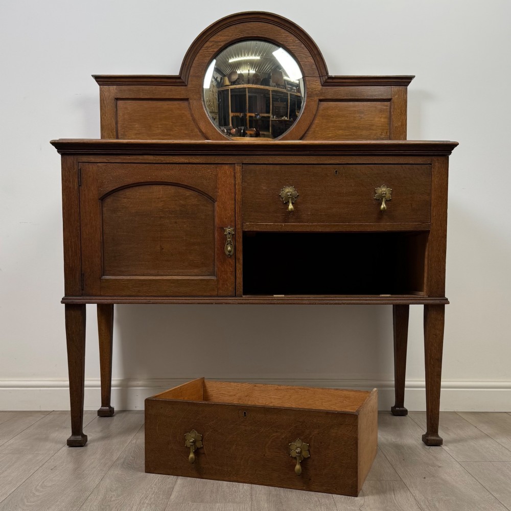 An Edwardian oak sideboard featuring a raised gallery back with a central circular mirror, a left cupboard door, two right drawers, and brass drop handles. View XI