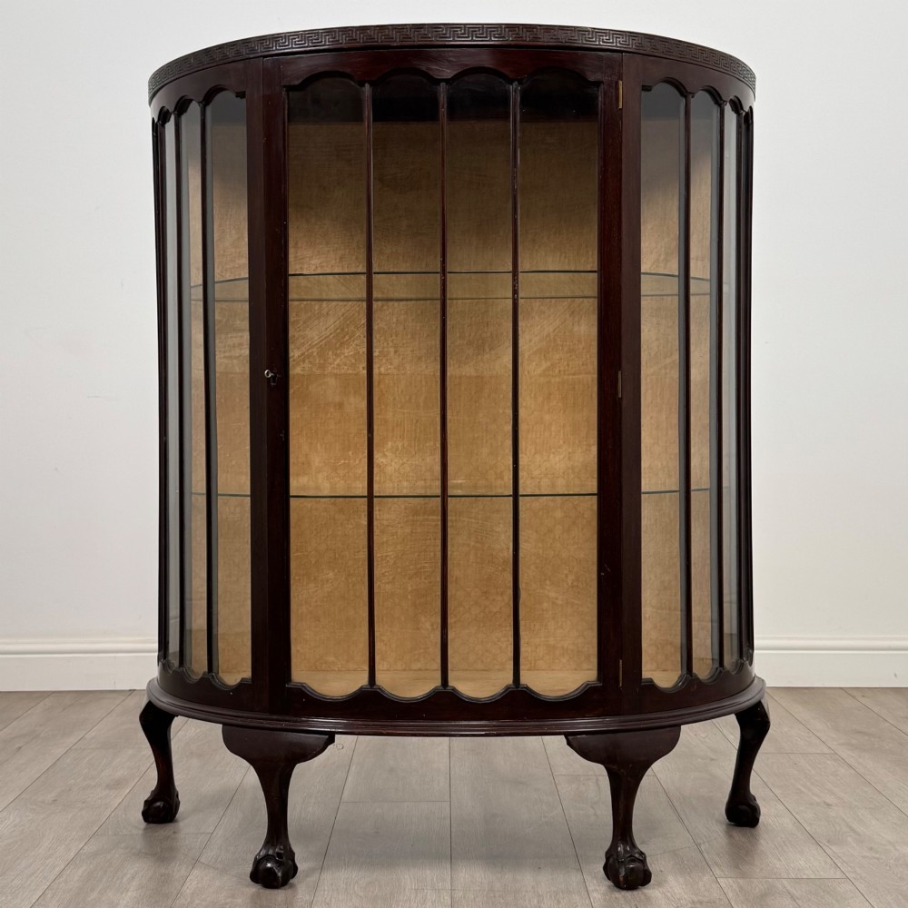 An Edwardian dark mahogany demi-lune display cabinet featuring glazed curved panels, a carved Greek key frieze, glass internal shelves, and cabriole legs with ball-and-claw feet.