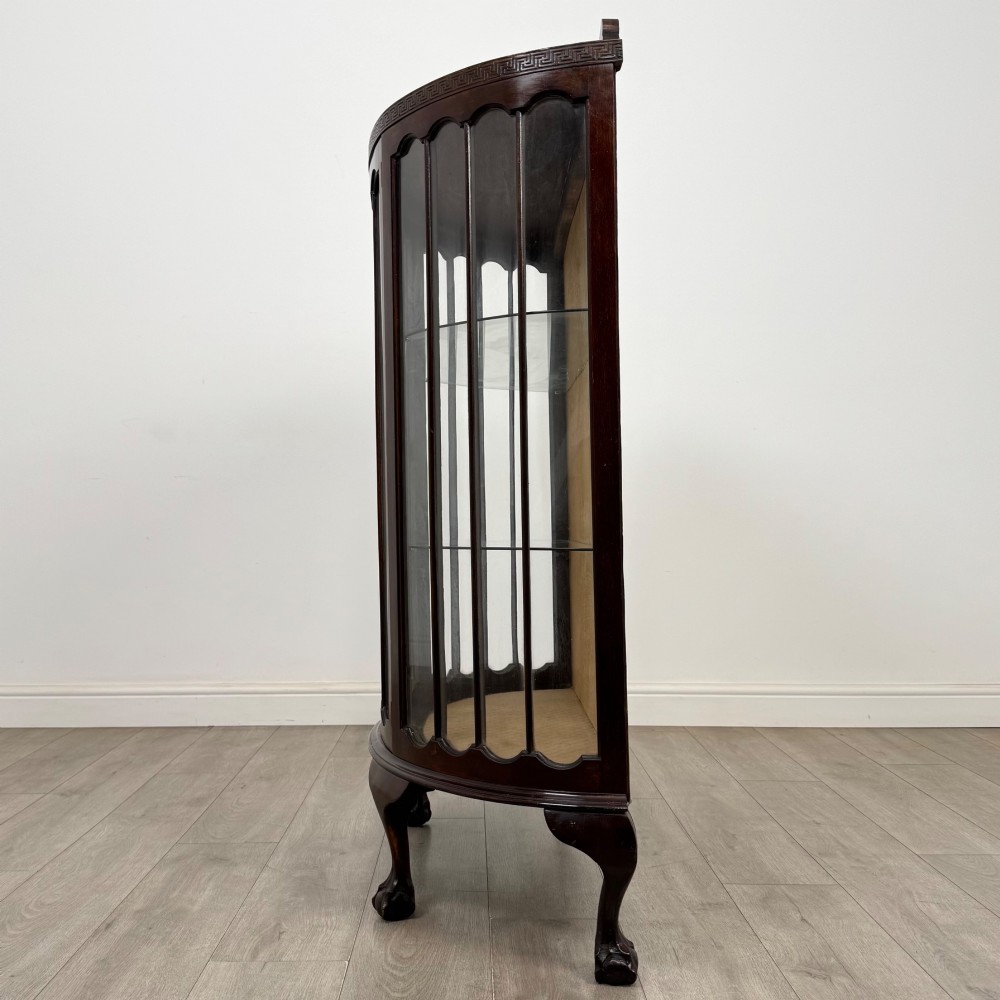 An Edwardian dark mahogany demi-lune display cabinet featuring glazed curved panels, a carved Greek key frieze, glass internal shelves, and cabriole legs with ball-and-claw feet. View IV