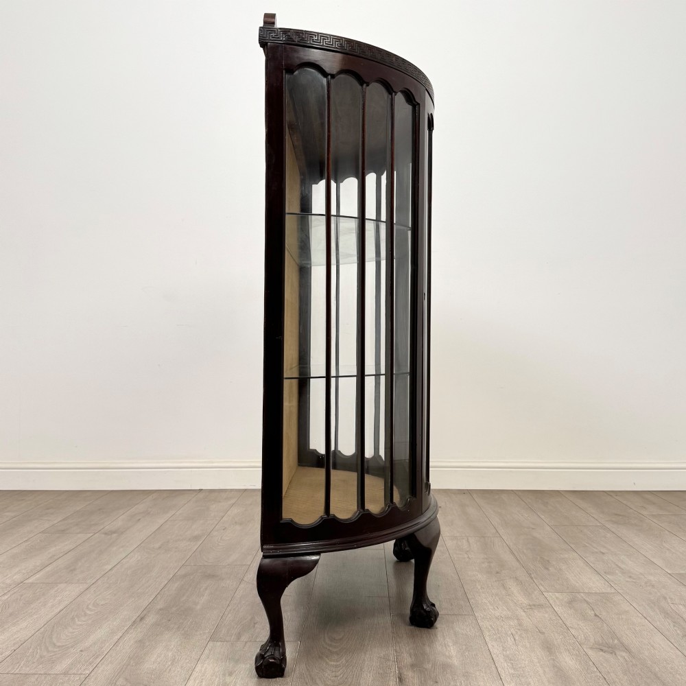 An Edwardian dark mahogany demi-lune display cabinet featuring glazed curved panels, a carved Greek key frieze, glass internal shelves, and cabriole legs with ball-and-claw feet. View VI
