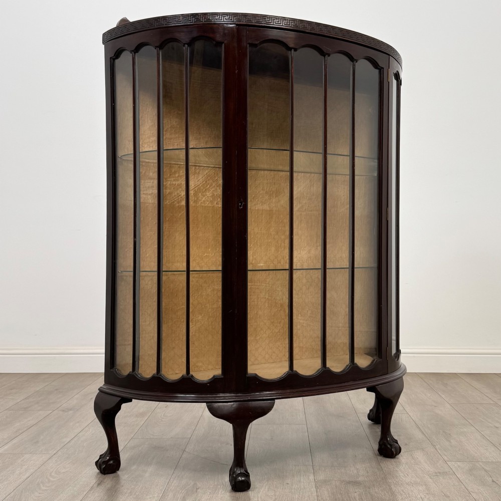 An Edwardian dark mahogany demi-lune display cabinet featuring glazed curved panels, a carved Greek key frieze, glass internal shelves, and cabriole legs with ball-and-claw feet. View VII