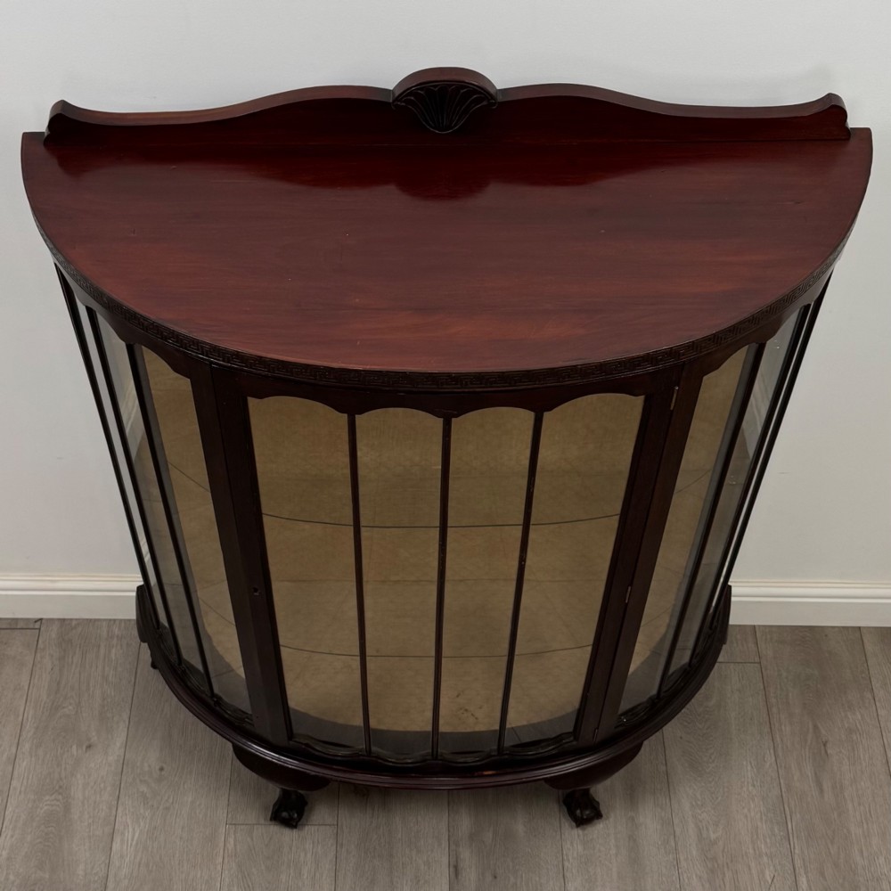 An Edwardian dark mahogany demi-lune display cabinet featuring glazed curved panels, a carved Greek key frieze, glass internal shelves, and cabriole legs with ball-and-claw feet. View VIII