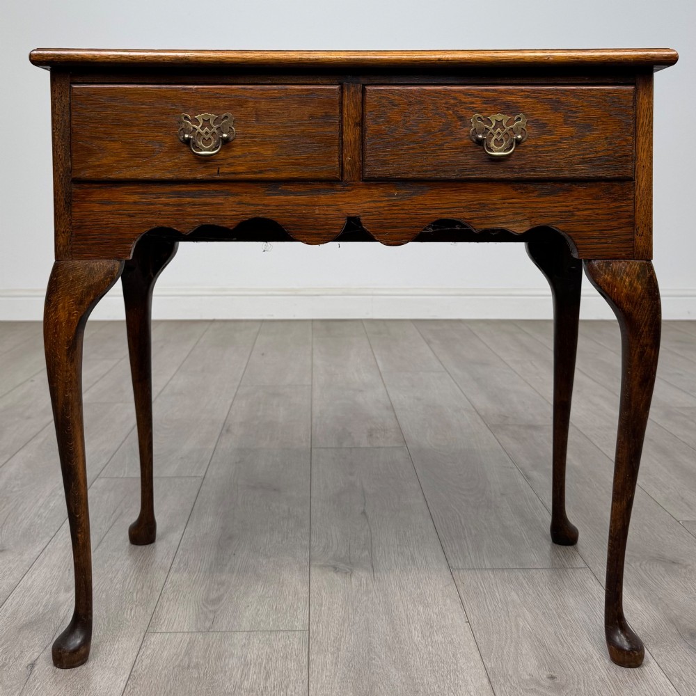 An Edwardian oak side table featuring two front drawers with brass drop handles, a scalloped apron, and slender cabriole legs.