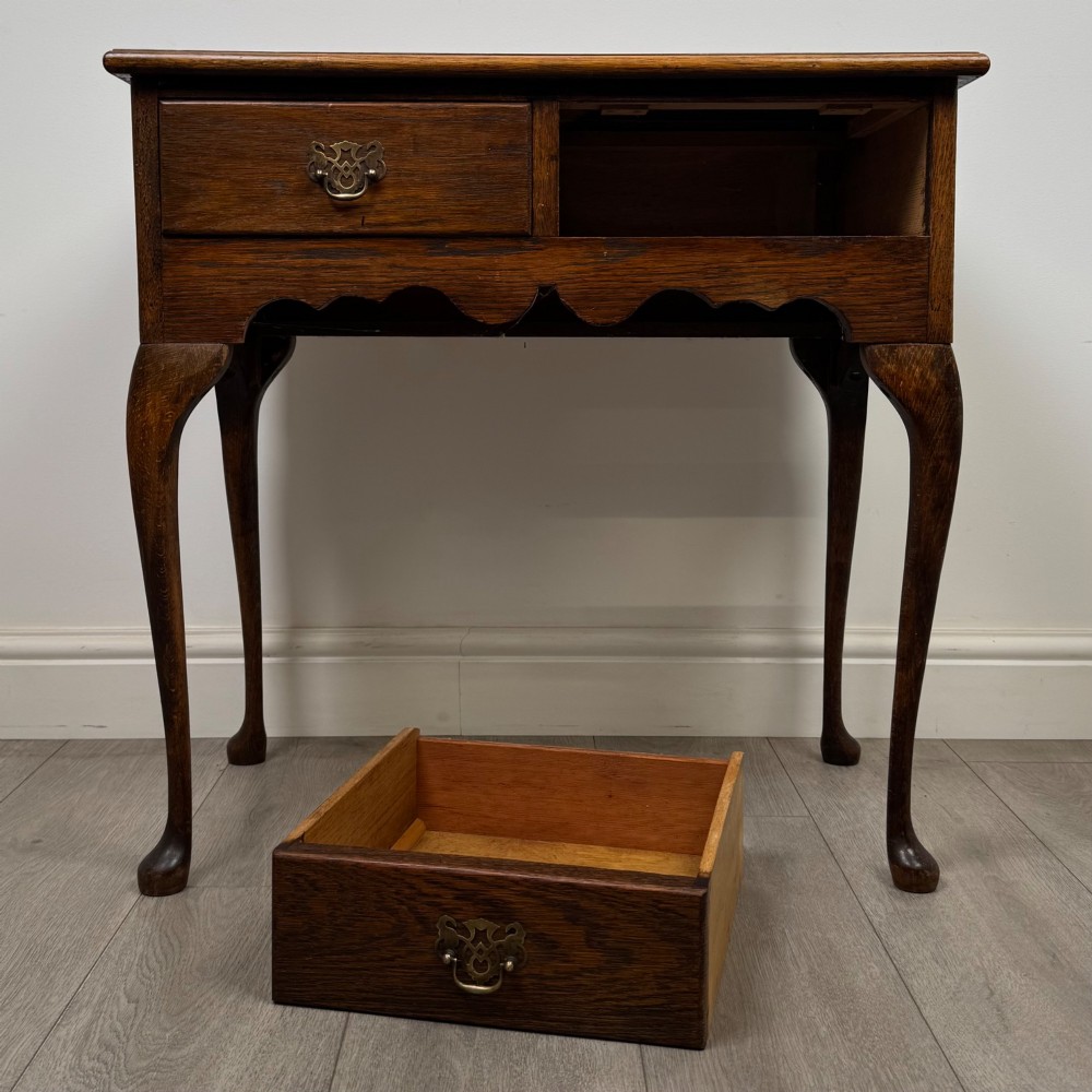 An Edwardian oak side table featuring two front drawers with brass drop handles, a scalloped apron, and slender cabriole legs. View X