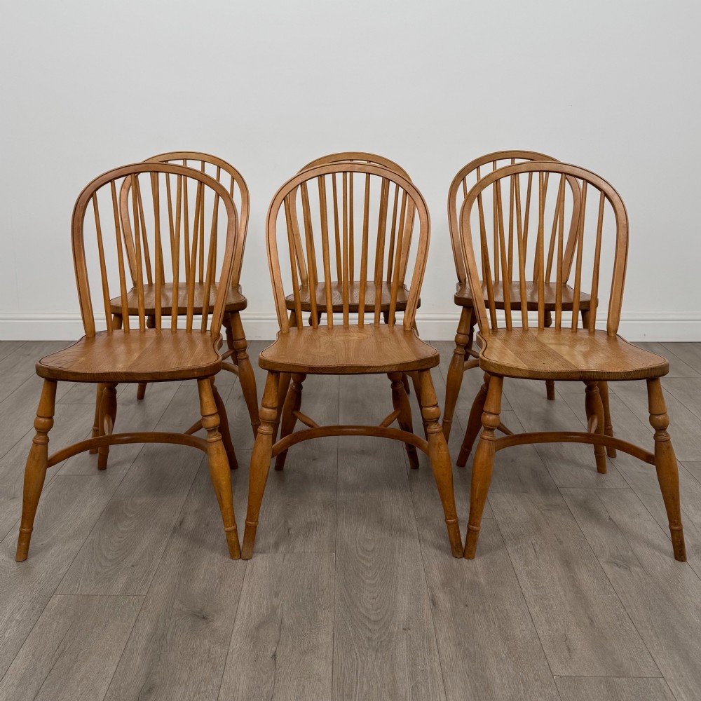 A set of six light warm brown ash Windsor dining chairs featuring hoop backs, turned spindles, shaped wooden seats, and turned legs with stretchers.