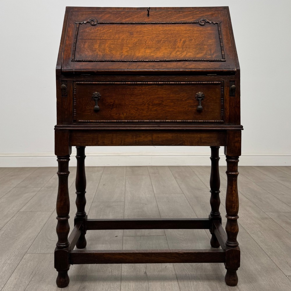 A dark oak Jacobean style bureau featuring a slanted drop-front lid, geometric moulding, a single drawer with teardrop handles, and turned legs with low stretchers.