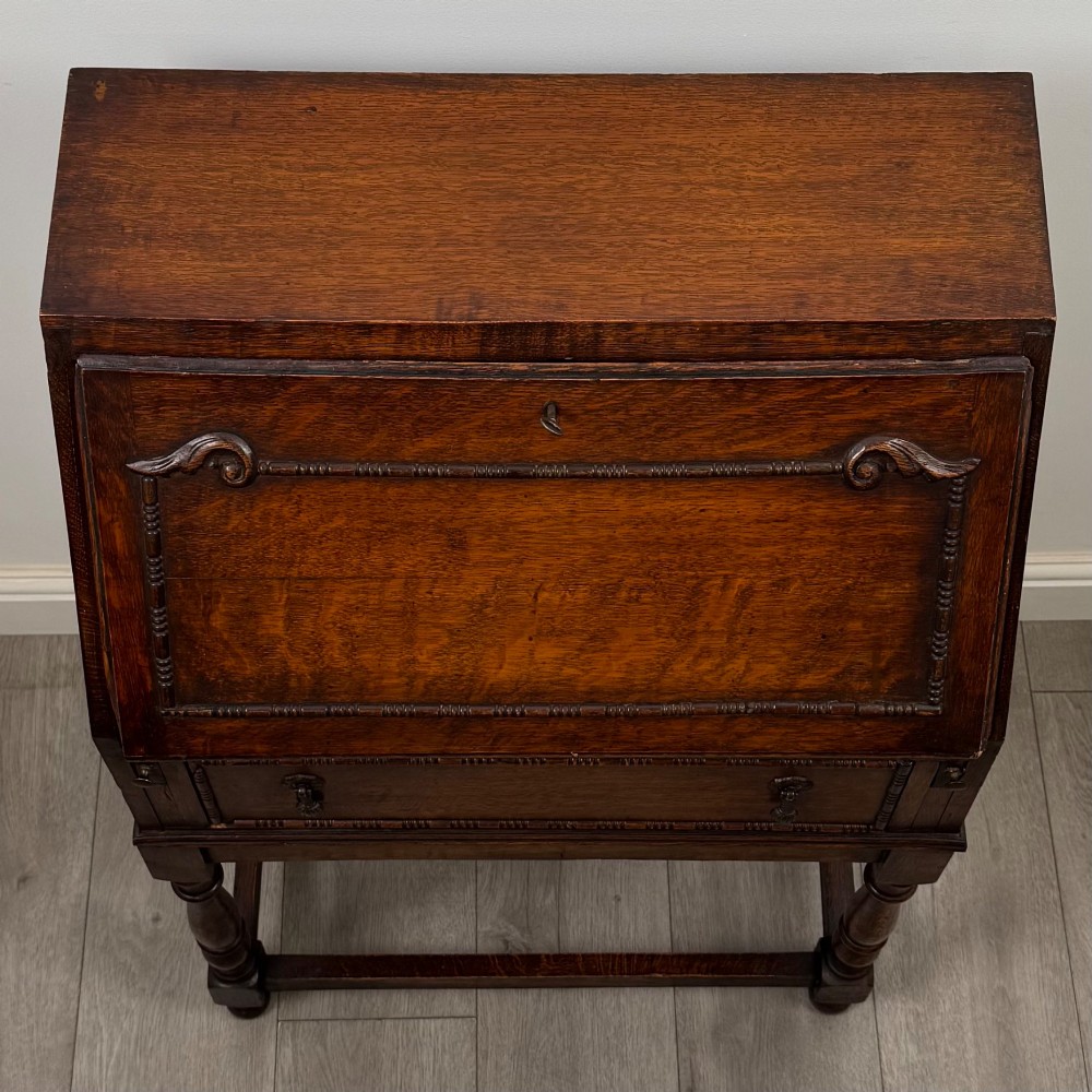 A dark oak Jacobean style bureau featuring a slanted drop-front lid, geometric moulding, a single drawer with teardrop handles, and turned legs with low stretchers. View X