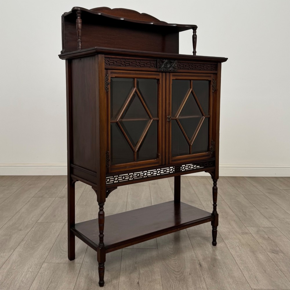 An Edwardian mahogany display cabinet featuring twin diamond-glazed doors, an upper shelf gallery, decorative fretwork, turned support legs, and an undertier. View IX