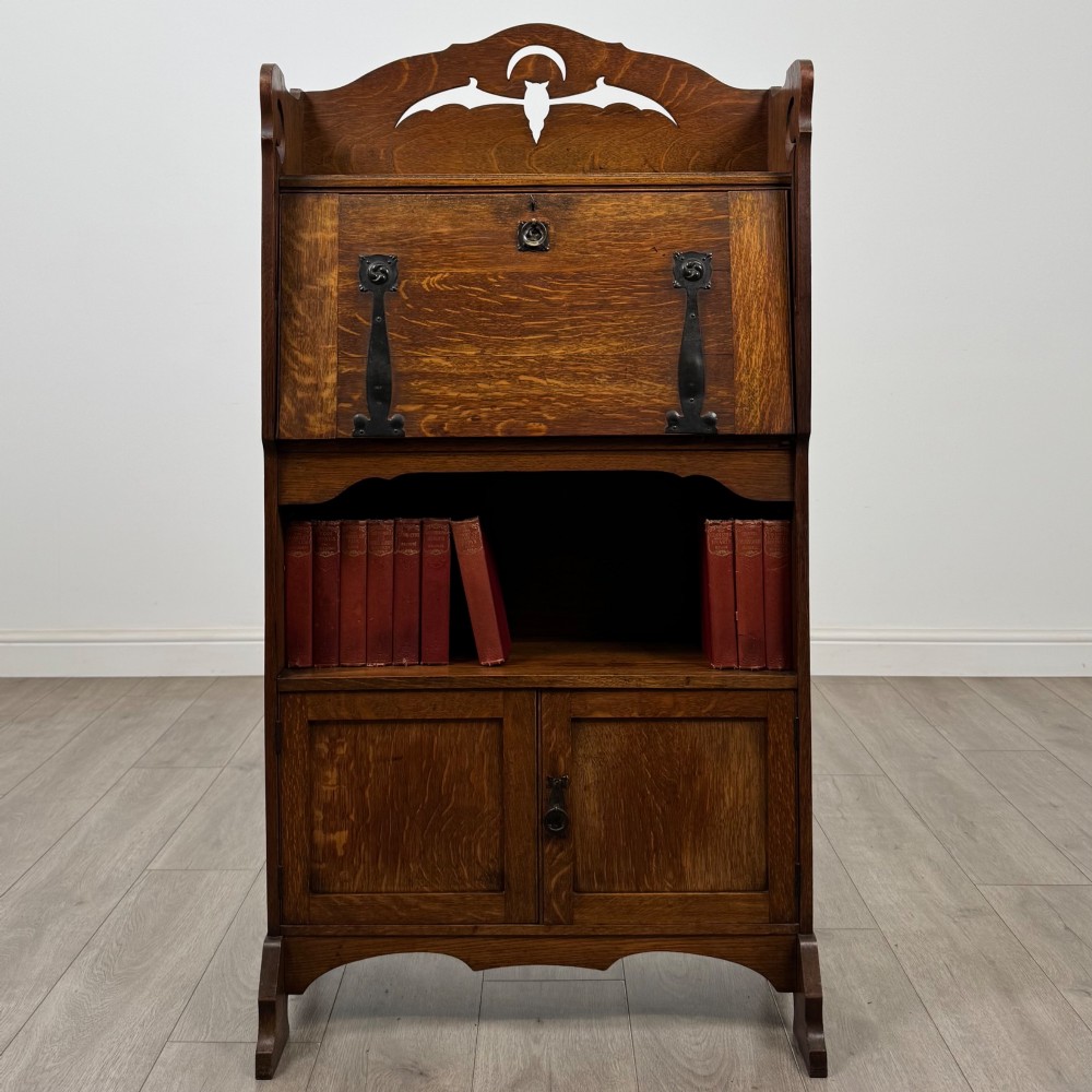 An antique Arts and Crafts oak writing bureau featuring a slant drop-front desk, a pierced cresting top rail, dark iron strap hardware, an open shelf, and lower double doors.