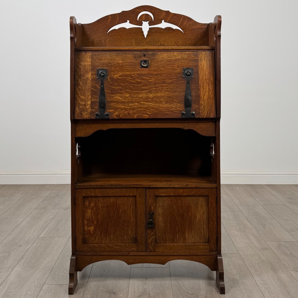 An antique Arts and Crafts oak writing bureau featuring a slant drop-front desk, a pierced cresting top rail, dark iron strap hardware, an open shelf, and lower double doors. View II