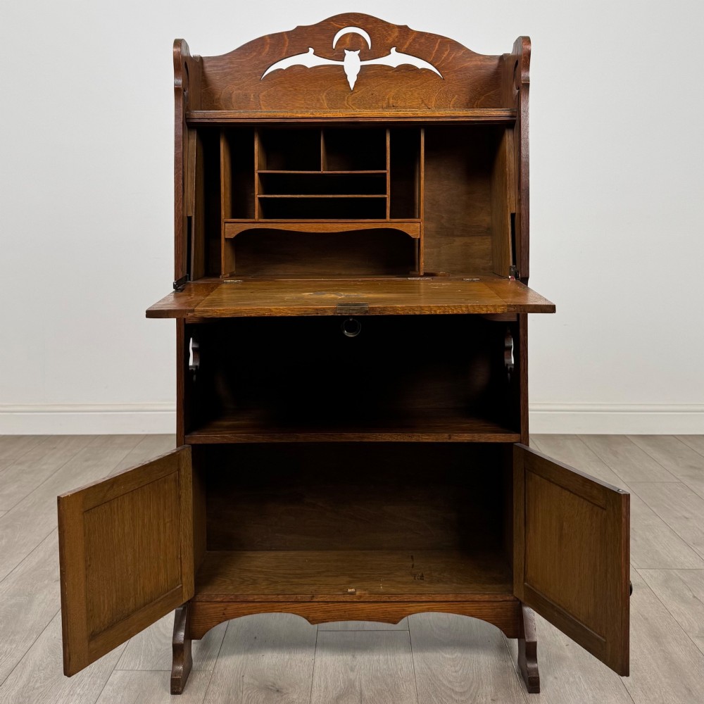 An antique Arts and Crafts oak writing bureau featuring a slant drop-front desk, a pierced cresting top rail, dark iron strap hardware, an open shelf, and lower double doors. View III
